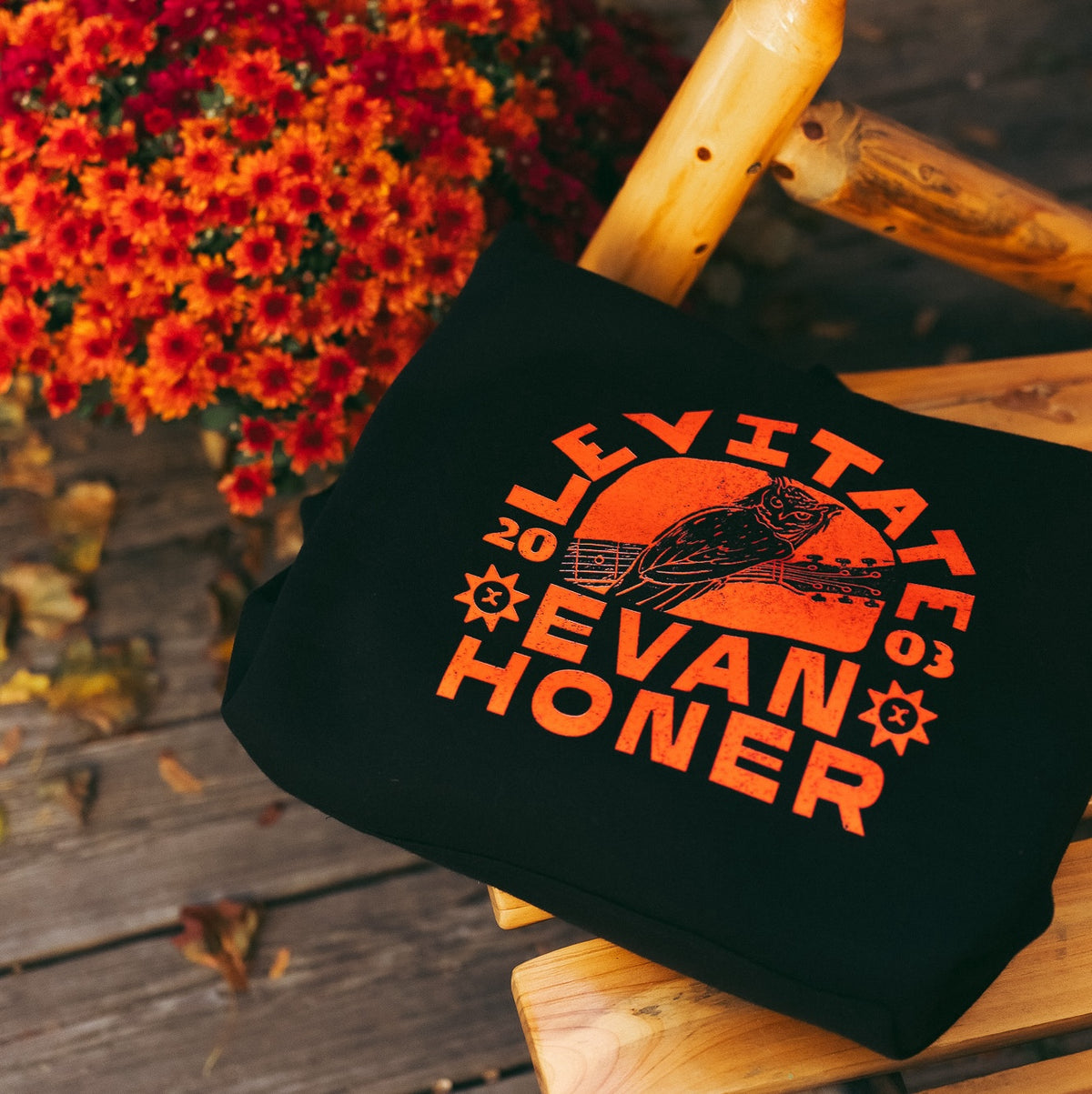 Levitate x Evan Honer Hoodie, front: folded black hoodie with the orange owl and guitar print on a wooden chair beside orange mums