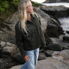 A woman with long blonde hair stands on a rocky shoreline wearing an olive green button-up jacket and light blue jeans, looking off-camera toward the water.