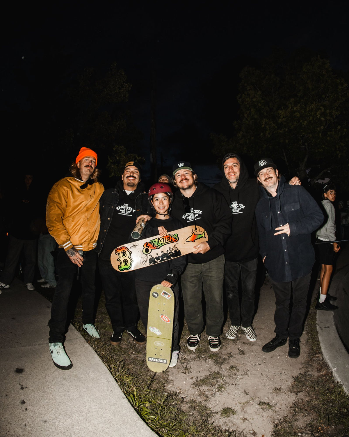 The Elovaters and skaters posing together at night after the Shark Belly Classic, holding up a skateboard