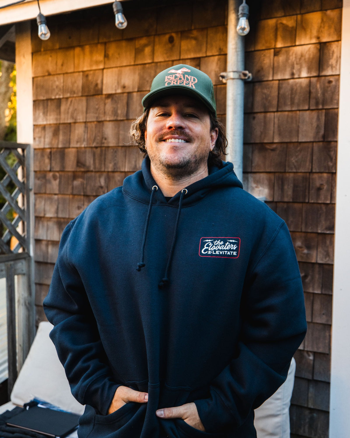 Member of The Elovaters smiling in a navy Levitate x Elovaters hoodie and trucker hat on the Backyard patio