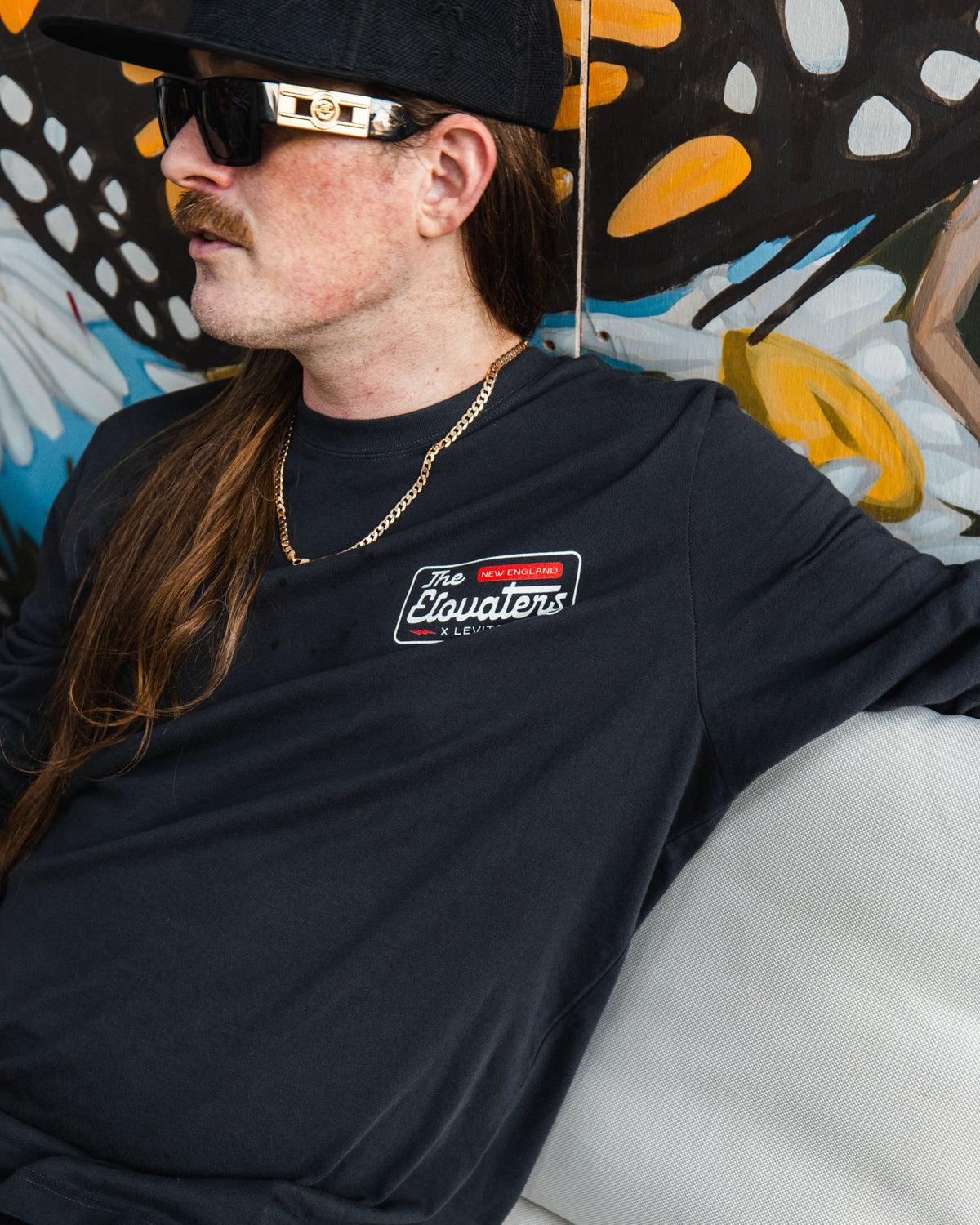 Member of The Elovaters in sunglasses and the collab tee in front of a Backyard mural