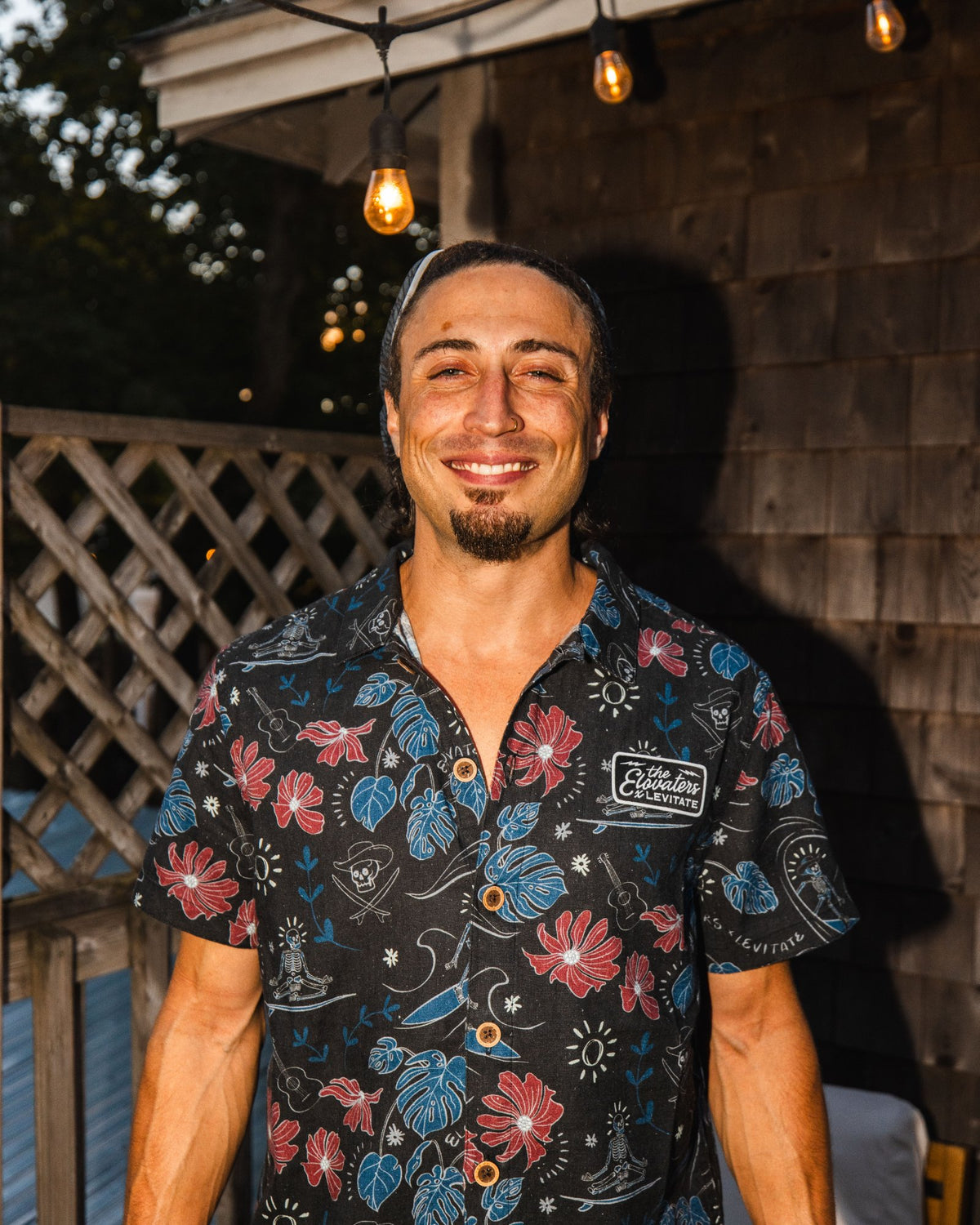 Member of The Elovaters smiling in the floral Levitate x Elovaters Beach Cantina Shirt under Backyard string lights