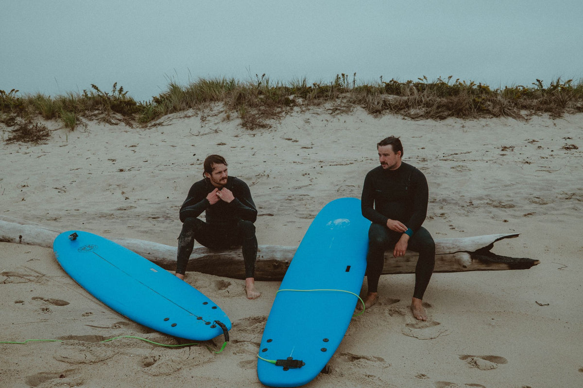 Evan Honer and a friend in wetsuits sitting on a driftwood log beside two blue surfboards on the beach