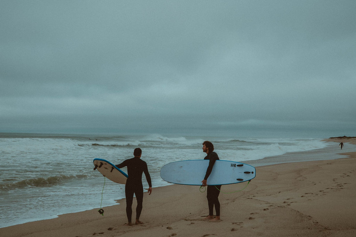Evan Honer and a friend in wetsuits carrying blue surfboards toward the water on a gray Nantucket morning