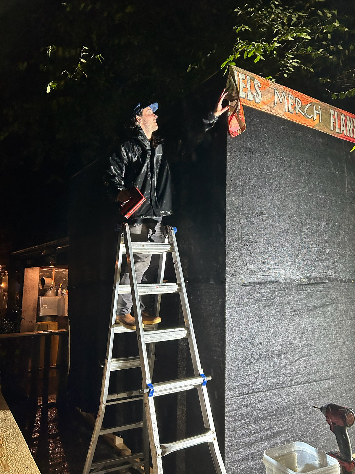 Crew member on a ladder hanging a sign at night during the Flannel Jam build at Cisco Brewers